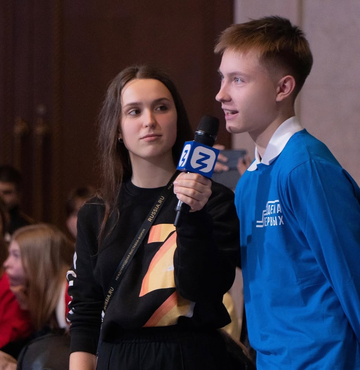 A young man in a blue shirt speaks into a microphone that an interviewer holds for him at a public event.